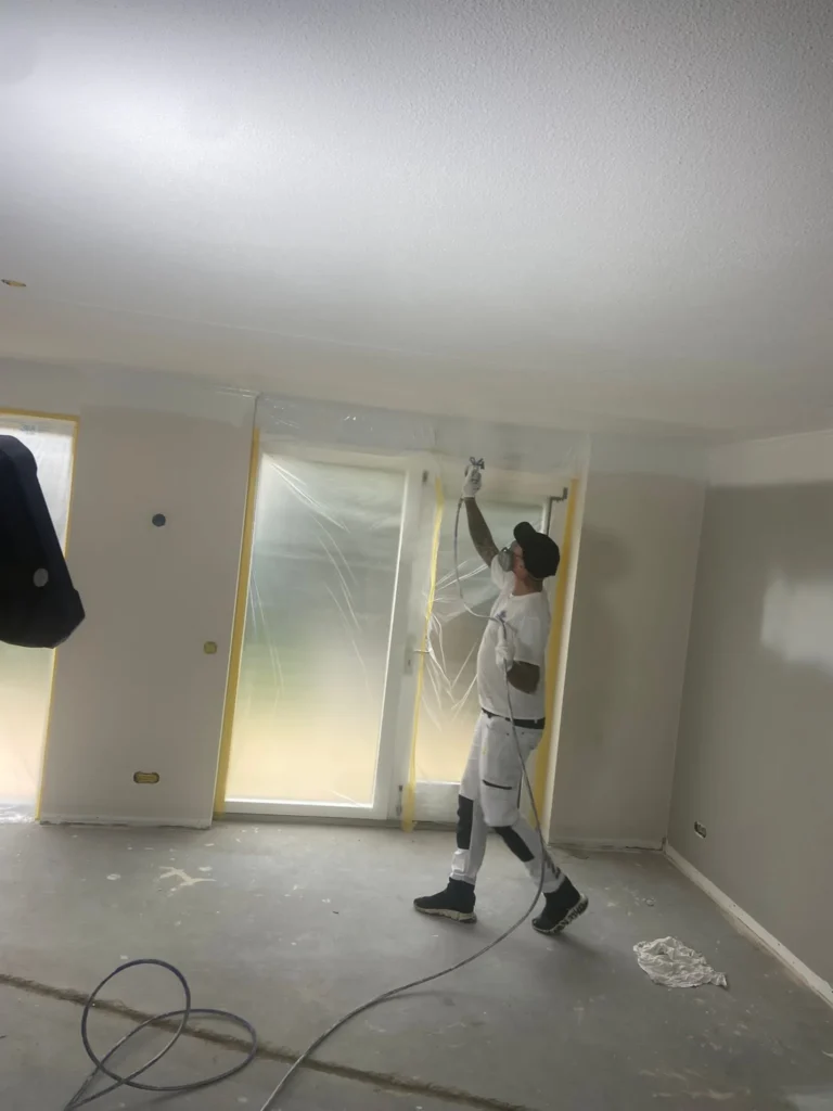 Schilder met spuitmasker spuit het plafond wit in een lege ruimte met afgeplakte ramen in Castricum